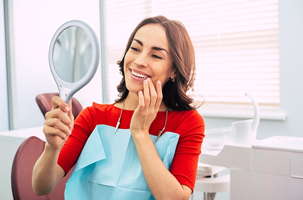 Woman in orange-red shirt and blue bib admiring smile in handheld mirror