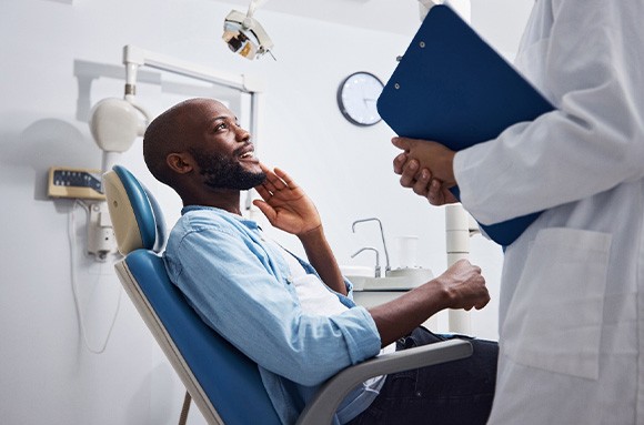 Man in blue shirt in dental chair talking to dentist with hand held to jaw