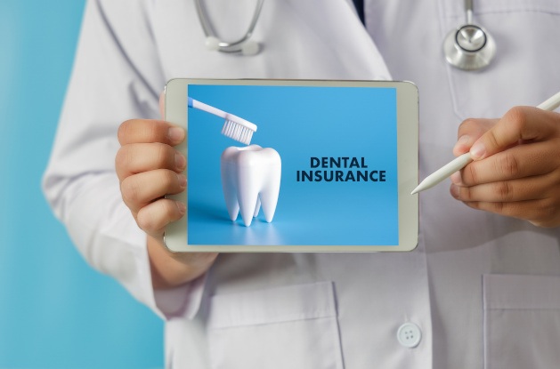 Dentist in white coat holding iPad with “Dental Insurance” showing next to a tooth and toothbrush with light blue background